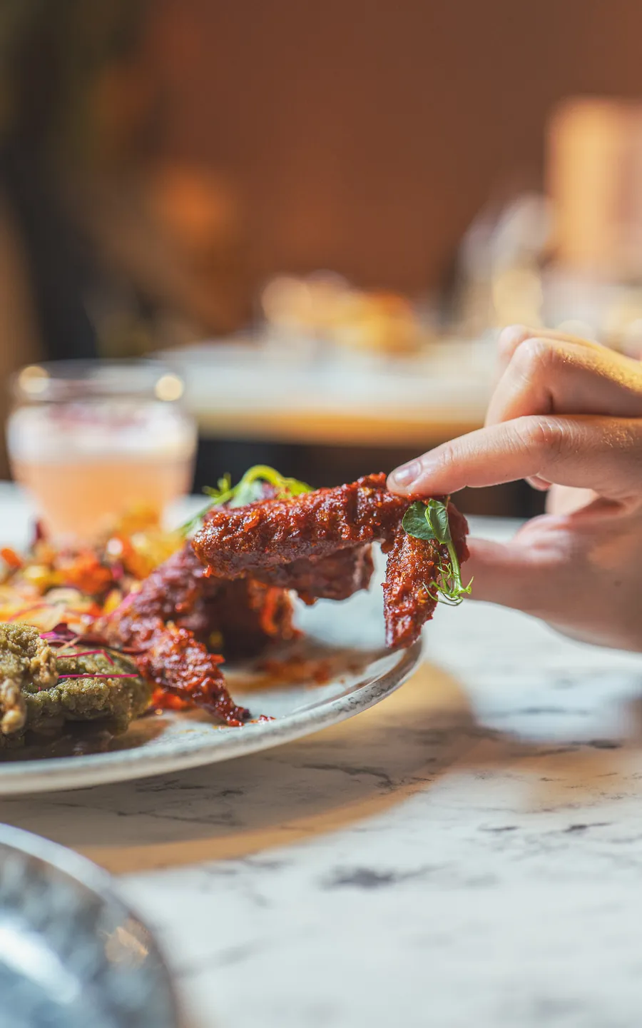 A hand lifting a glazed Bombay chicken wing off the plate