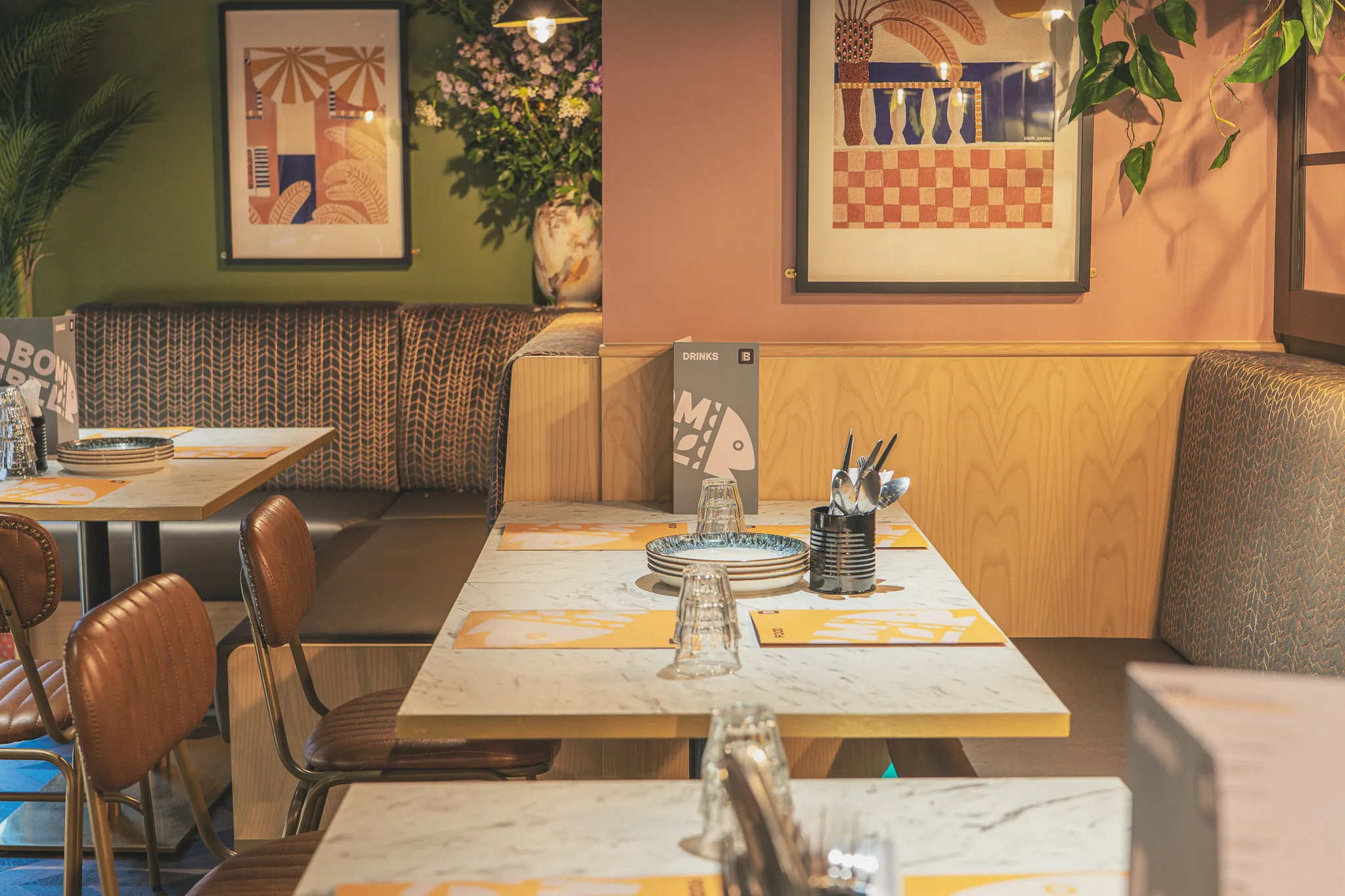 A blush-pink corner with framed prints, a woven booth and tables laid for dinner