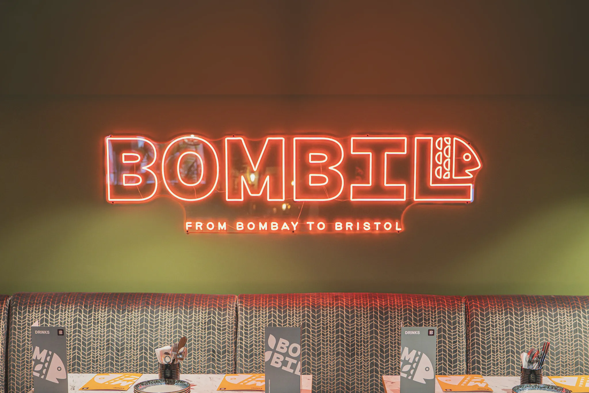 Red neon reading Bombil, from Bombay to Bristol, above patterned booths on a bottle-green wall